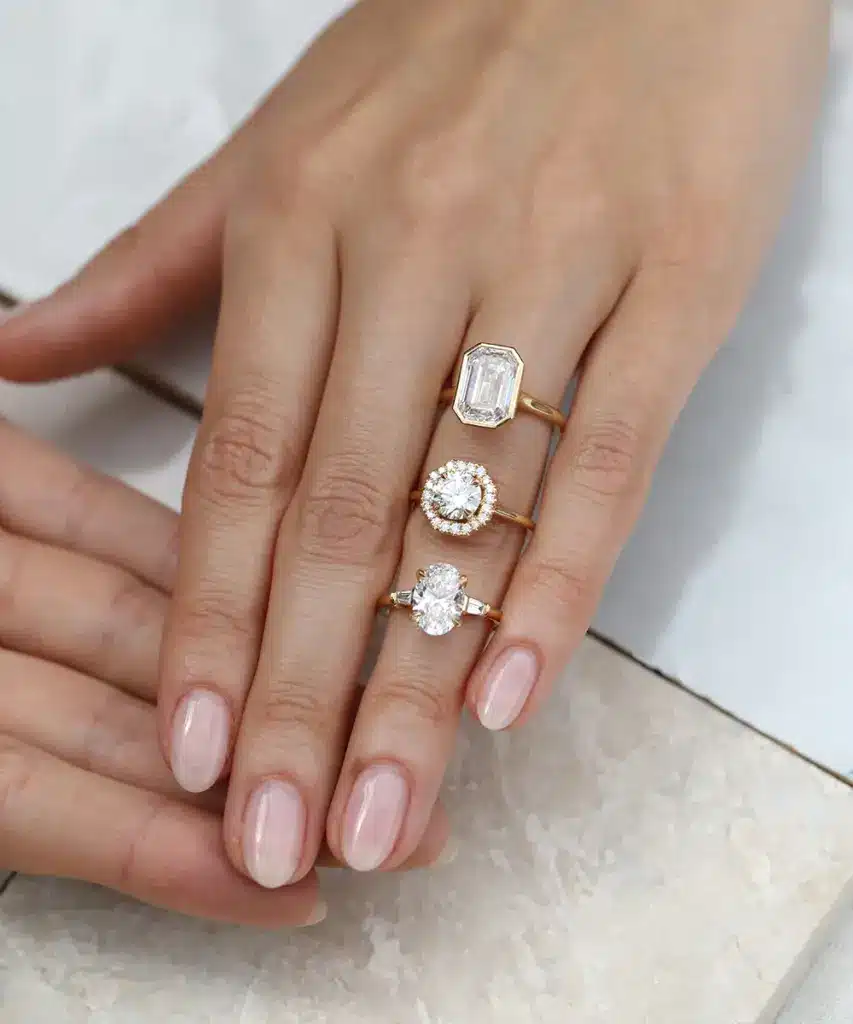 AE0A6714 Hand wearing three elegant gold diamond rings on polished nails, displayed on a marble surface for a luxurious fashion statement. | Sky Rye Design Hand wearing three elegant gold diamond rings on polished nails, displayed on a marble surface for a luxurious fashion statement.