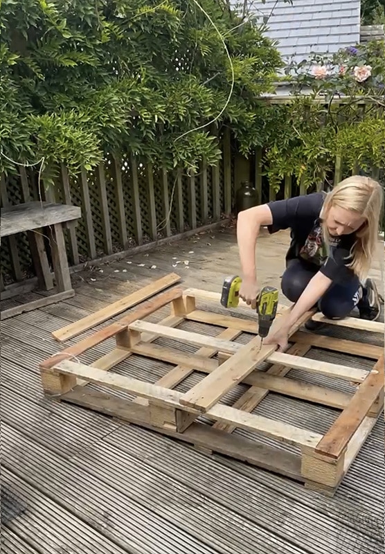 Adding-new-slats- Person using a power drill to assemble a wooden pallet on a sunny deck, surrounded by green foliage. | Sky Rye Design Person using a power drill to assemble a wooden pallet on a sunny deck, surrounded by green foliage.