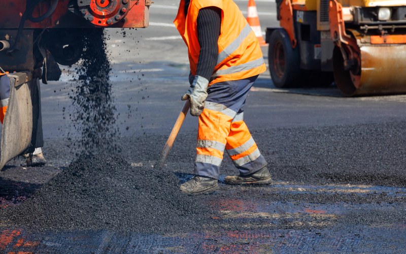 Road worker in orange safety gear laying asphalt with an industrial machine on a sunny day.