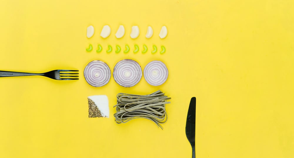 Neatly arranged pasta ingredients on a yellow background with garlic, onion, green peppers, salt, pepper, and a fork and knife.