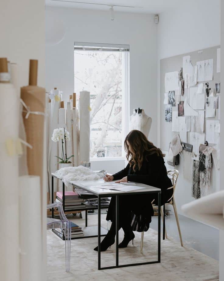Antoinette Fusillo not only designs but meticulously oversees every step of the bridal couture journey ensuring each of our delightful brides receives the wedding gown of their dreams_ Fashion designer sketching in a studio surrounded by fabrics and inspiration boards with natural light streaming through a large window. | Sky Rye Design Fashion designer sketching in a studio surrounded by fabrics and inspiration boards with natural light streaming through a large window.