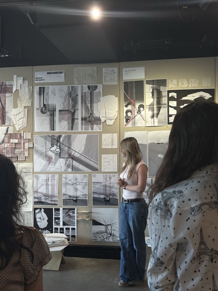 Architecture Woman presenting architectural sketches and models on display board in a gallery setting, observed by two individuals. | Sky Rye Design Woman presenting architectural sketches and models on display board in a gallery setting, observed by two individuals.