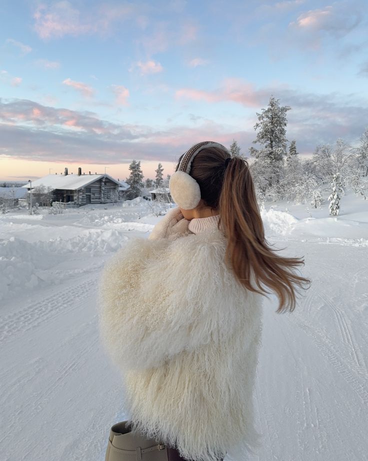 Arrived in a magical Christmas wonderland with my family 💫 happy to be back auroracollectionresort Woman in cozy fur coat and earmuffs enjoying a snowy landscape at sunset, with rustic cabin and pine trees in the background. | Sky Rye Design Woman in cozy fur coat and earmuffs enjoying a snowy landscape at sunset, with rustic cabin and pine trees in the background.