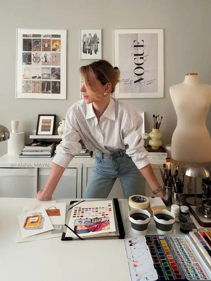 Artist in a studio with colorful palettes and art supplies, planning designs. Fashion magazine covers adorn the wall in the background.