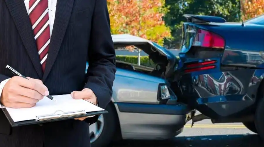 Man in suit with clipboard assessing a rear-end car accident damage on a sunny day, focusing on insurance and claims evaluation.
