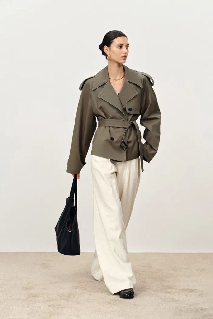 Beatrice Short Trench Coat in Cotton Twill Elegant woman in an olive trench coat and white trousers, holding a black bag, showcasing modern fashion against a neutral background. | Sky Rye Design Elegant woman in an olive trench coat and white trousers, holding a black bag, showcasing modern fashion against a neutral background.
