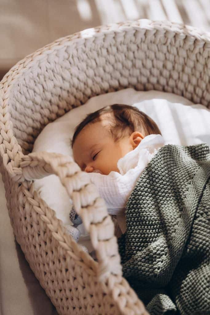 Berço Moisés Sleeping baby in a cozy knit bassinet, wrapped in a soft green blanket. Peaceful, serene scene of newborn resting in sunlight. | Sky Rye Design Sleeping baby in a cozy knit bassinet, wrapped in a soft green blanket. Peaceful, serene scene of newborn resting in sunlight.
