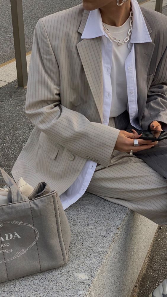 Blazer Outfit Person in pinstripe suit sitting on a bench, holding a phone, with a designer tote bag nearby. Stylish and modern fashion scene. | Sky Rye Design Person in pinstripe suit sitting on a bench, holding a phone, with a designer tote bag nearby. Stylish and modern fashion scene.