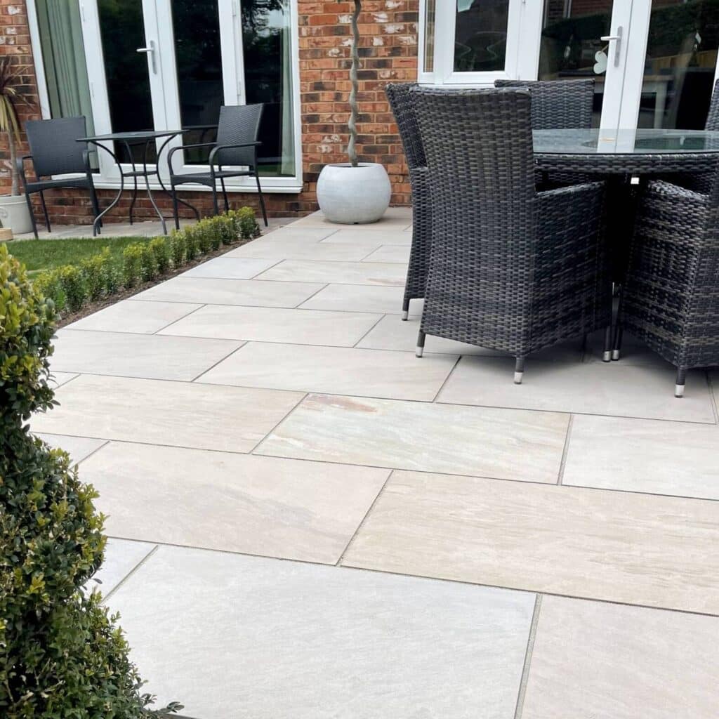 Camel-Buff-in-situ-1-1574-x-1574 Modern patio with gray paving stones, outdoor dining set, black wicker chairs, and potted plants by a brick house and glass doors. | Sky Rye Design Modern patio with gray paving stones, outdoor dining set, black wicker chairs, and potted plants by a brick house and glass doors.