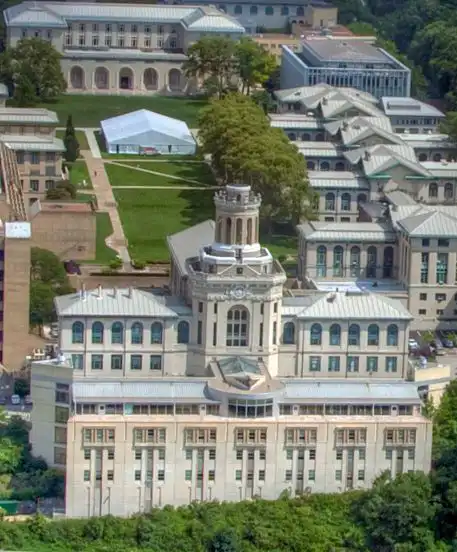 Aerial view of a historic university building with a clock tower, surrounded by lush greenery and academic buildings on a sunny day.