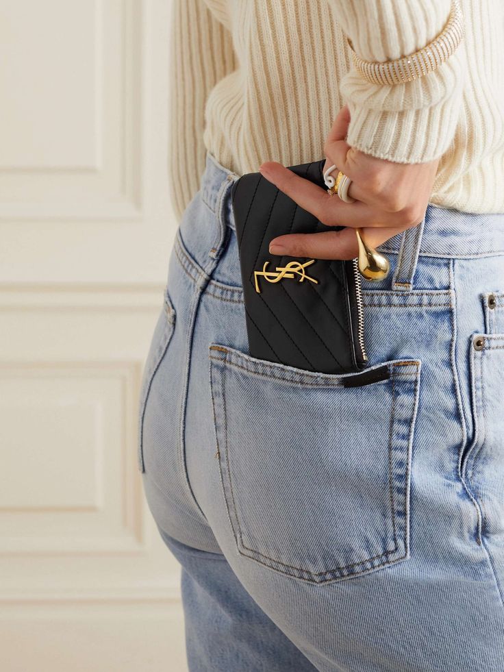 Person in jeans placing a black quilted cardholder with gold logo into back pocket, wearing a beige sweater and stylish accessories.