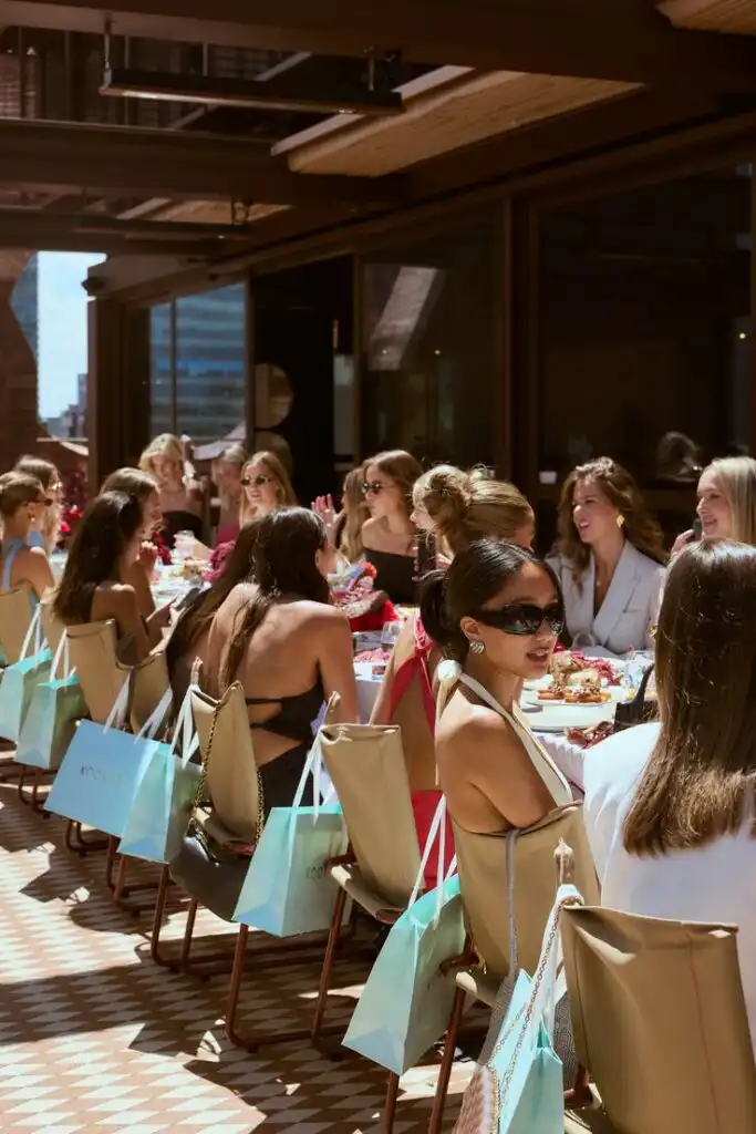 Celebrating 33 Years of Kookai Group of women enjoying an outdoor brunch with gift bags on a sunny day, surrounded by modern architecture. | Sky Rye Design Group of women enjoying an outdoor brunch with gift bags on a sunny day, surrounded by modern architecture.