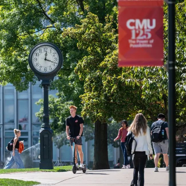 Centers Institutes Campus scene with students walking and scootering near a clock and CMU banner, surrounded by trees and sunlight. | Sky Rye Design Campus scene with students walking and scootering near a clock and CMU banner, surrounded by trees and sunlight.
