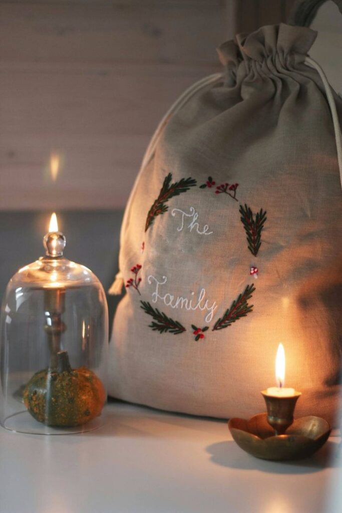 Christmas Drawstring Linen Sack With Hand Embroidered Family Name_ Reusable Embroidered Linen Pouch _ Christmas Gifts Sack - Etsy UK Cozy holiday decor with a candle-lit setting, featuring a fabric bag labeled The Family adorned with festive embroidery. | Sky Rye Design Cozy holiday decor with a candle-lit setting, featuring a fabric bag labeled The Family adorned with festive embroidery.