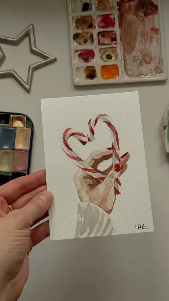 Christmas watercolor card tutorial Watercolor painting of a hand holding candy canes shaped as a heart, surrounded by art supplies on a table. | Sky Rye Design Watercolor painting of a hand holding candy canes shaped as a heart, surrounded by art supplies on a table.
