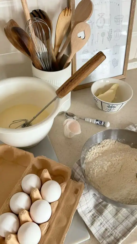 Baking essentials on countertop: whisk, eggs, flour, butter, and wooden utensils. Perfect setup for a cozy baking day.