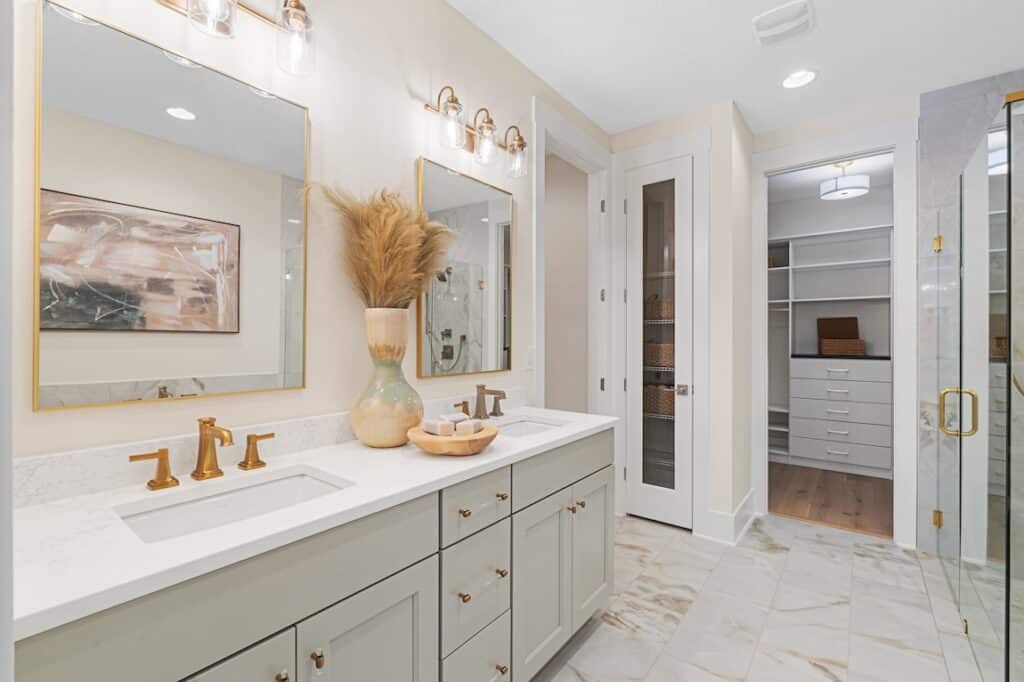 Crosswinds-at-Nocatee_Woodlawn-Elite_Master-Bath_03 Modern bathroom with double vanity, marble countertop, gold fixtures, and storage space, leading to a walk-in closet. | Sky Rye Design Modern bathroom with double vanity, marble countertop, gold fixtures, and storage space, leading to a walk-in closet.