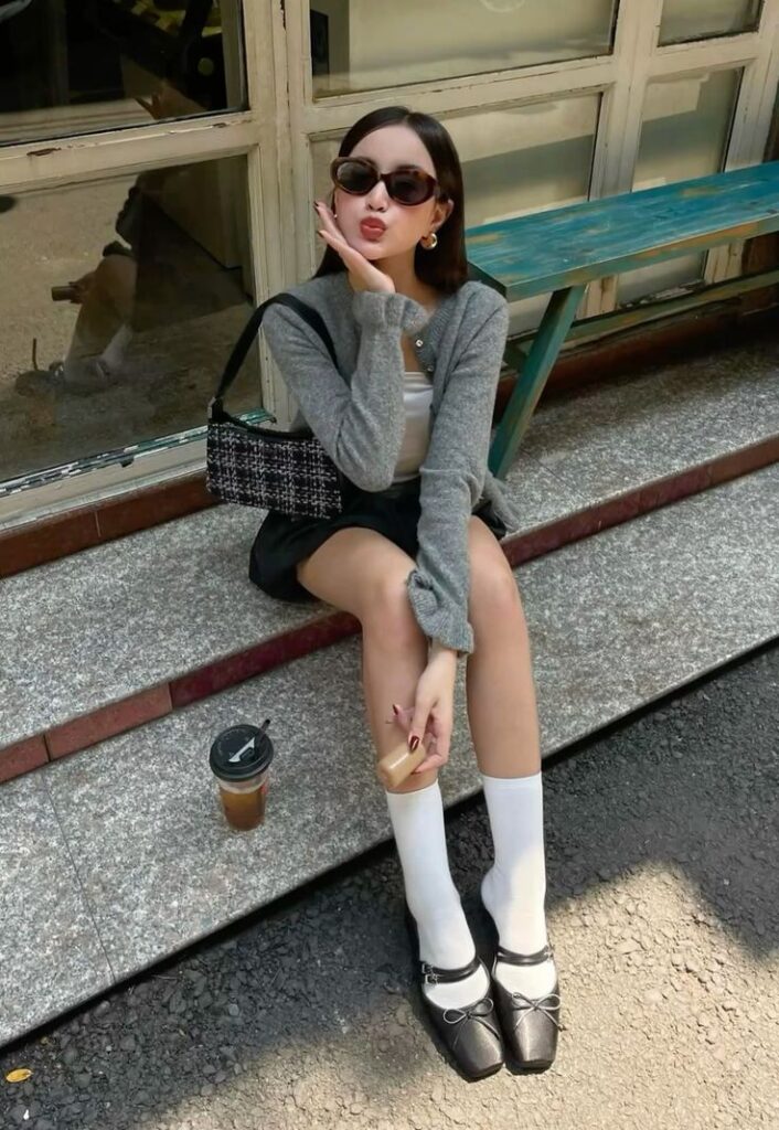 DC🍎 Stylish woman in sunglasses sitting outside, wearing a gray cardigan, black skirt, and Mary Jane shoes, holding a drink. | Sky Rye Design Stylish woman in sunglasses sitting outside, wearing a gray cardigan, black skirt, and Mary Jane shoes, holding a drink.
