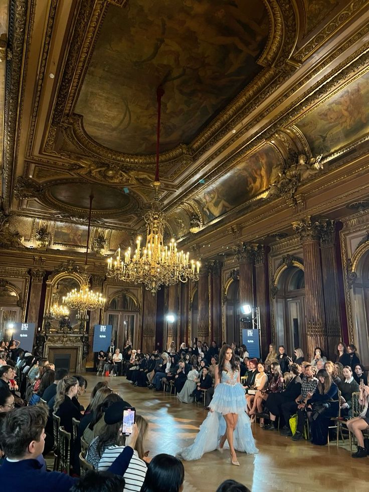 Desiree Deschampsstyle fashion Model in blue gown walks runway in opulent hall with chandeliers, surrounded by an audience, showcasing stunning fashion design. | Sky Rye Design Model in blue gown walks runway in opulent hall with chandeliers, surrounded by an audience, showcasing stunning fashion design.