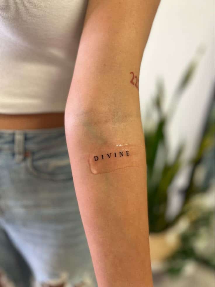 Close-up of a person's arm with a bandage featuring DIVINE text. Arm shows a small, red tattoo in background. Casual summer attire.