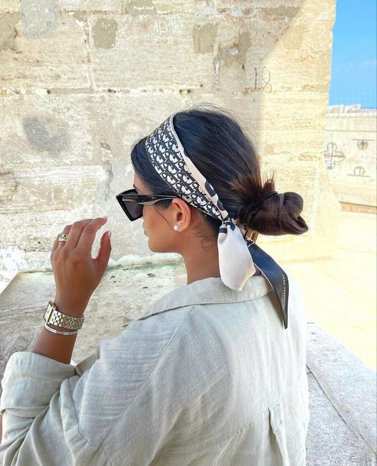 Dior _ summer look Woman with sunglasses and patterned headscarf relaxes outdoors, wearing a light blouse and elegant jewelry, against a stone background. | Sky Rye Design Woman with sunglasses and patterned headscarf relaxes outdoors, wearing a light blouse and elegant jewelry, against a stone background.