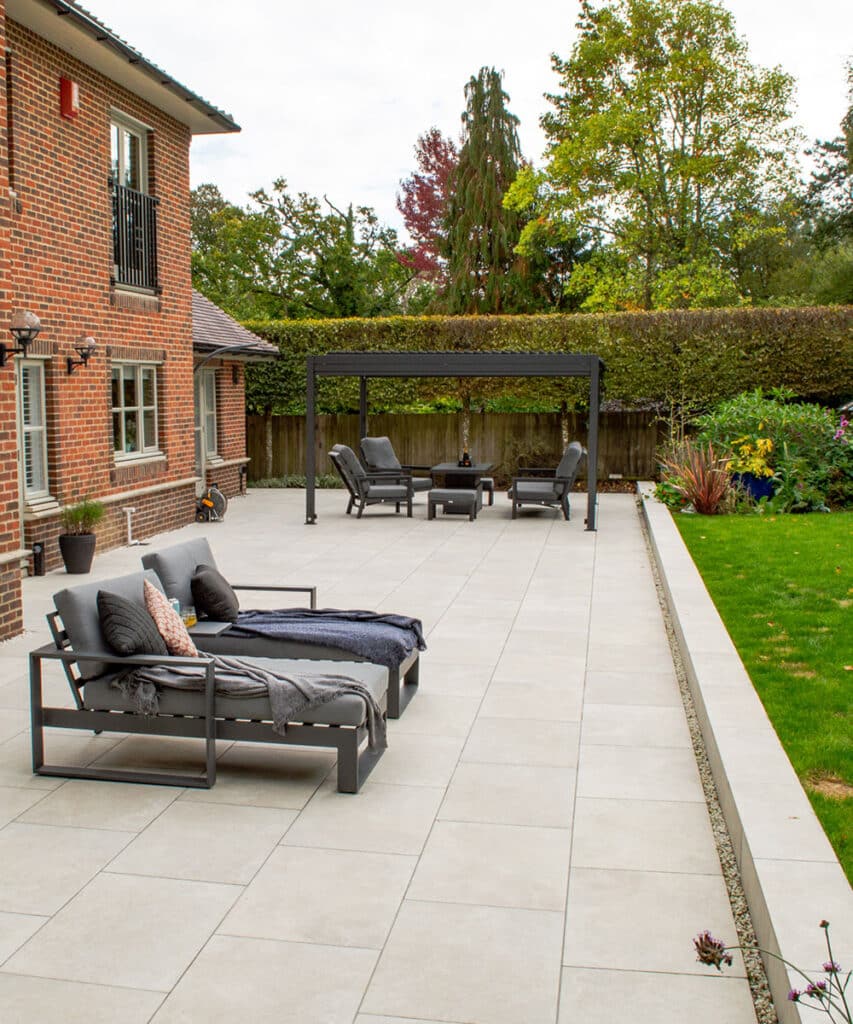Exbury-White-Italian-Porcelain-Paving-With-Tumbled-Edge-2 Spacious patio with modern outdoor seating, pergola, and lush garden, adjacent to a brick house, under a clear sky. Ideal for relaxation. | Sky Rye Design Spacious patio with modern outdoor seating, pergola, and lush garden, adjacent to a brick house, under a clear sky. Ideal for relaxation.