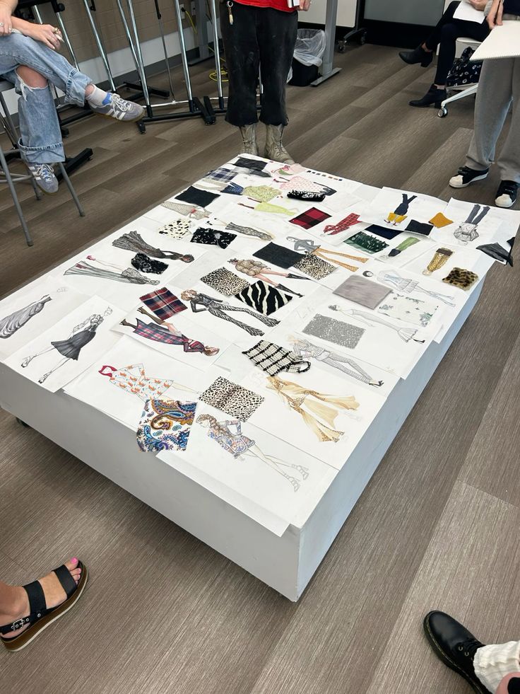 Fashion critique day Fashion design sketches and fabric swatches displayed on a table, surrounded by designers in a collaborative workspace. | Sky Rye Design Fashion design sketches and fabric swatches displayed on a table, surrounded by designers in a collaborative workspace.