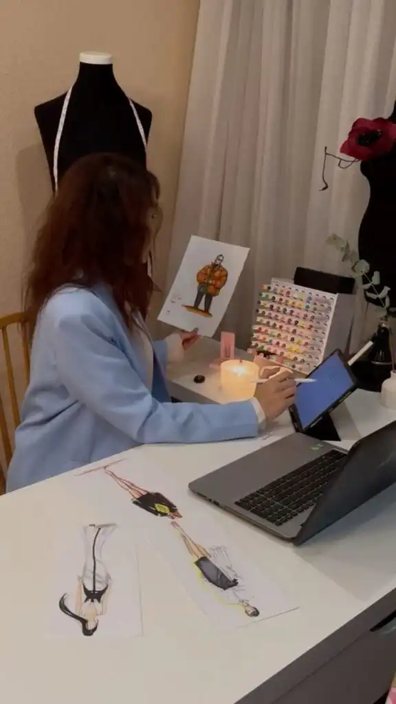 Fashion designer reviewing sketches at desk with laptop, drawing tools, and mannequin nearby; creative workspace setup. fashion line business plan