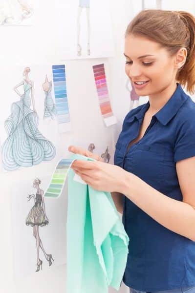 Fashion designer selecting fabric colors, smiling beside illustrated fashion sketches and color palettes pinned on a wall.