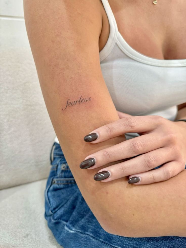 Close-up of a woman's arm with a fearless tattoo in elegant script, wearing a white tank top and blue jeans, dark polished nails visible.