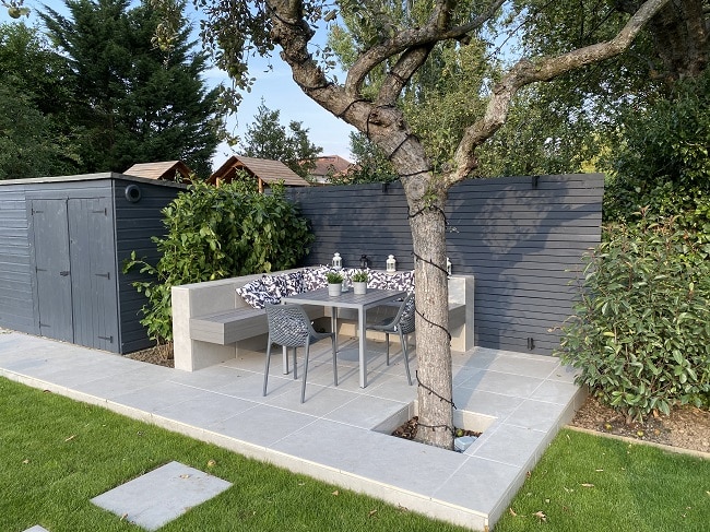 Florence-Grey-porcelain-patio-Pebble-Grey-Dark-Ash-Brushed-Limebok-Landscaping Modern backyard patio with a cozy dining area, featuring a tree centerpiece, gray fence, and garden shed for outdoor relaxation. | Sky Rye Design Modern backyard patio with a cozy dining area, featuring a tree centerpiece, gray fence, and garden shed for outdoor relaxation.