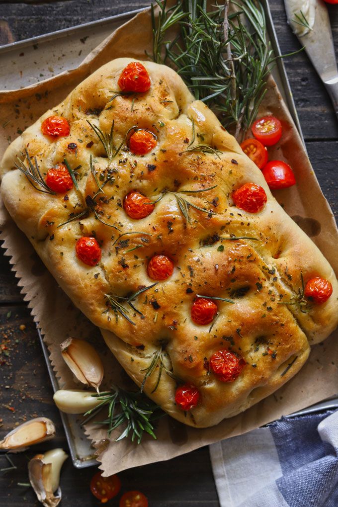 Focaccia Recipe Rosemary Garlic Bread - Fun FOOD Frolic Freshly baked focaccia with cherry tomatoes and rosemary on parchment paper, garnished with garlic cloves, on a rustic kitchen table. | Sky Rye Design Freshly baked focaccia with cherry tomatoes and rosemary on parchment paper, garnished with garlic cloves, on a rustic kitchen table.