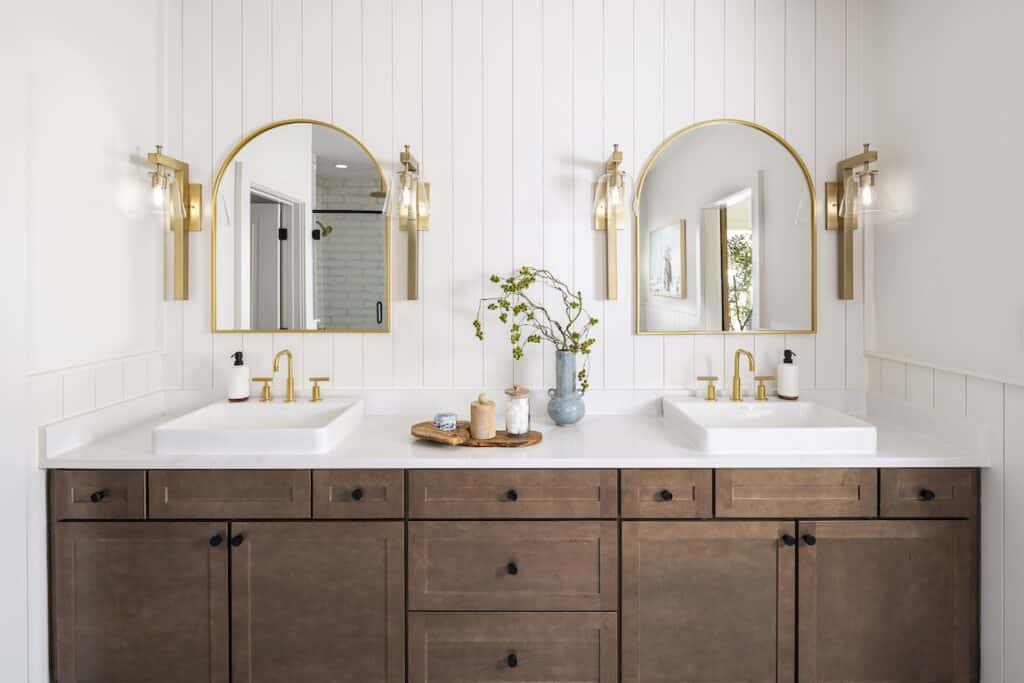 Freestone-Station_Hillside-Farmhouse-Model_Lot-69_Primary-Bath-1 Modern bathroom with dual sinks, gold fixtures, arched mirrors, and wooden cabinetry, featuring a sleek, minimalist design. | Sky Rye Design Modern bathroom with dual sinks, gold fixtures, arched mirrors, and wooden cabinetry, featuring a sleek, minimalist design.