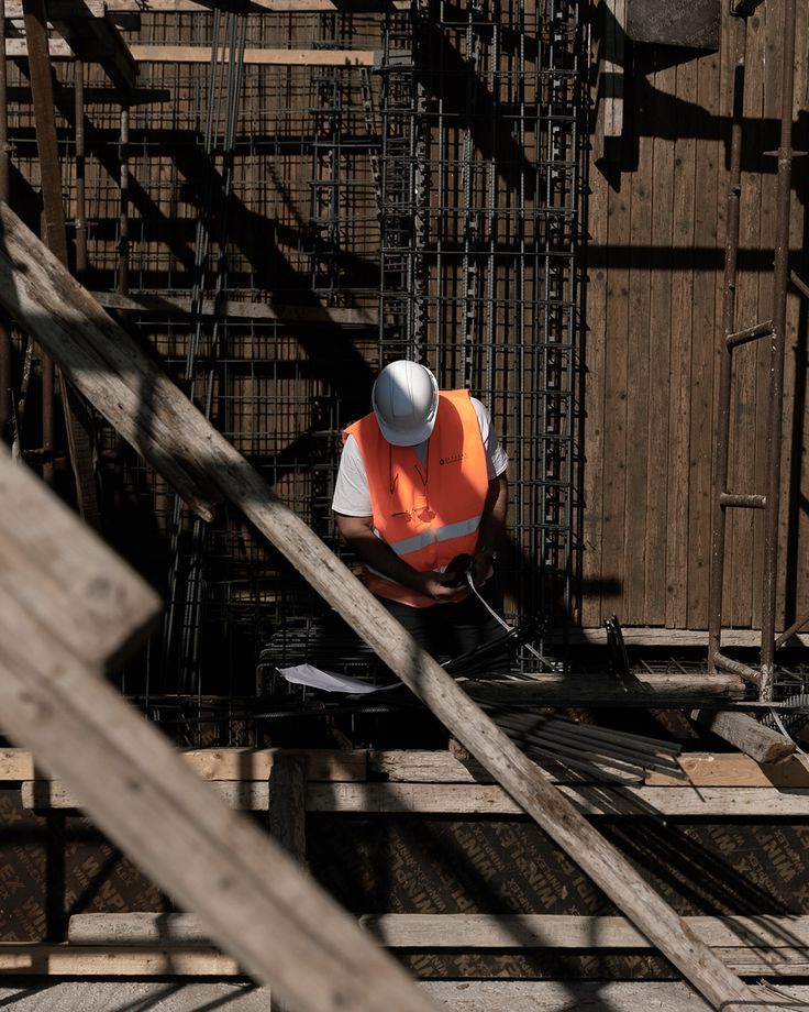 GROUNDWORK IN PROGRESS Construction worker in a safety vest and helmet inspects rebar framework at a building site. | Sky Rye Design Construction worker in a safety vest and helmet inspects rebar framework at a building site.