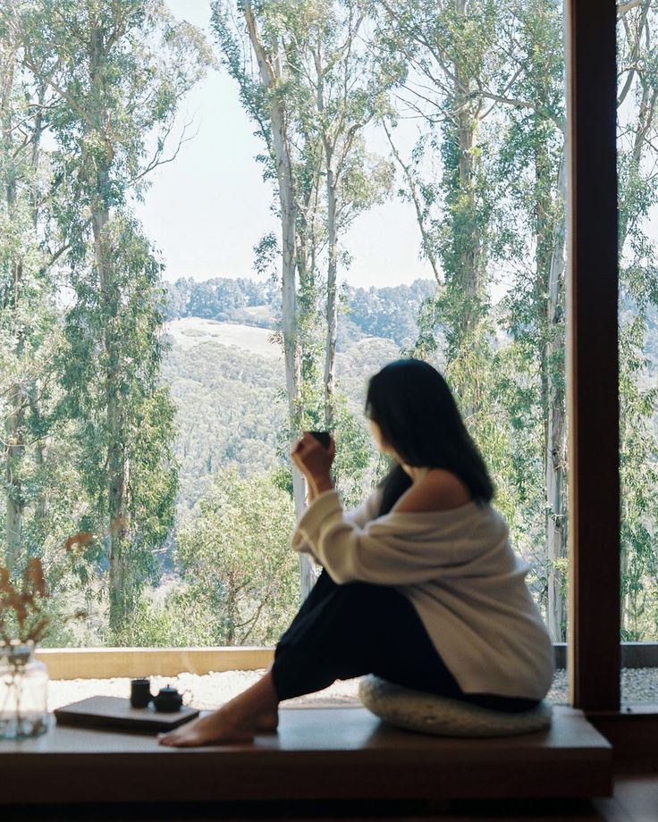 Gentleness simple rituals and prioritising rest 🕊️ Woman enjoys morning coffee by window, overlooking serene forest and hills. Peaceful and relaxing home setting. | Sky Rye Design Woman enjoys morning coffee by window, overlooking serene forest and hills. Peaceful and relaxing home setting.
