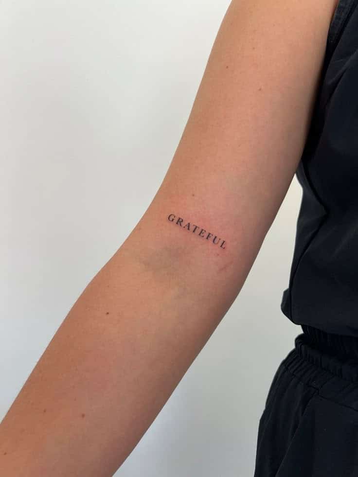 Arm with minimalist Grateful tattoo in black ink, seen against a white wall and black clothing.
