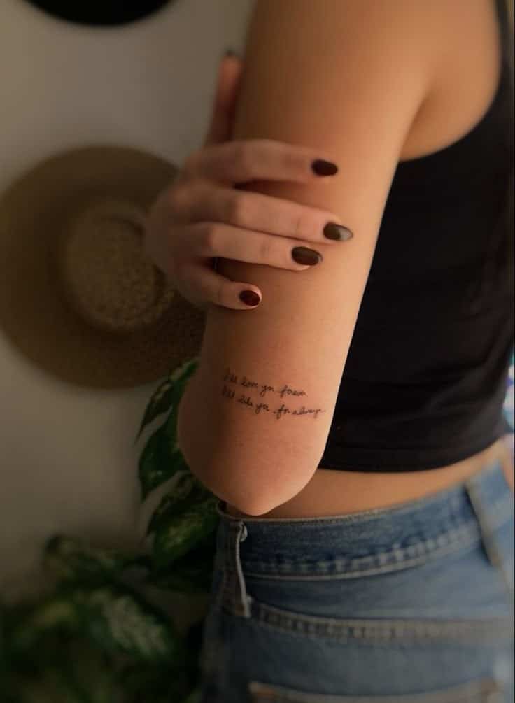 Woman with a meaningful script tattoo on her upper arm, wearing casual outfit, displaying unique nail polish, standing against a soft background.