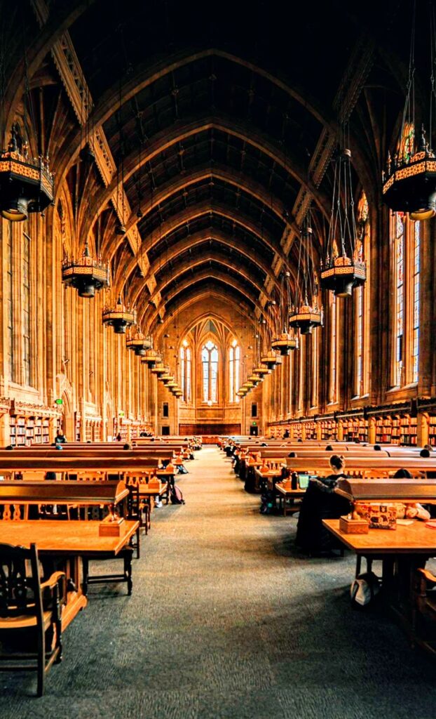 Harvard University 1 Gothic-style library interior with high vaulted ceilings, wooden tables, chandeliers, and large windows, creating a scholarly ambiance. | Sky Rye Design Gothic-style library interior with high vaulted ceilings, wooden tables, chandeliers, and large windows, creating a scholarly ambiance.