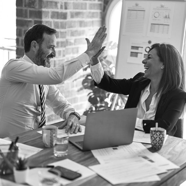 Business colleagues celebrating success with a high-five at a desk, smiling and surrounded by documents and a laptop. Black and white photo.