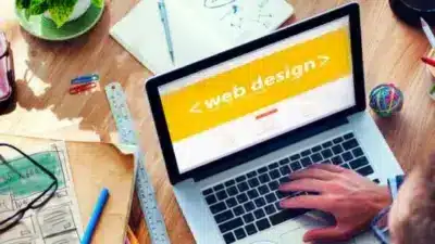 Person working on laptop with web design displayed, surrounded by desk items like notebooks, coffee, and potted plant.