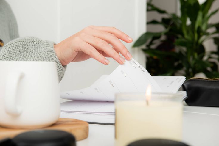 How To Be Consistent_ 7 Steps To Get You Motivated Again Person flipping through planner at cozy desk with candle and coffee mug, surrounded by plants, creating a relaxing work atmosphere. | Sky Rye Design Person flipping through planner at cozy desk with candle and coffee mug, surrounded by plants, creating a relaxing work atmosphere.