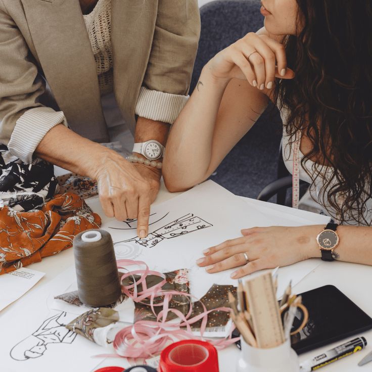 Two people discussing a fashion design sketch, surrounded by fabric swatches, ribbon, and sewing tools on a table.