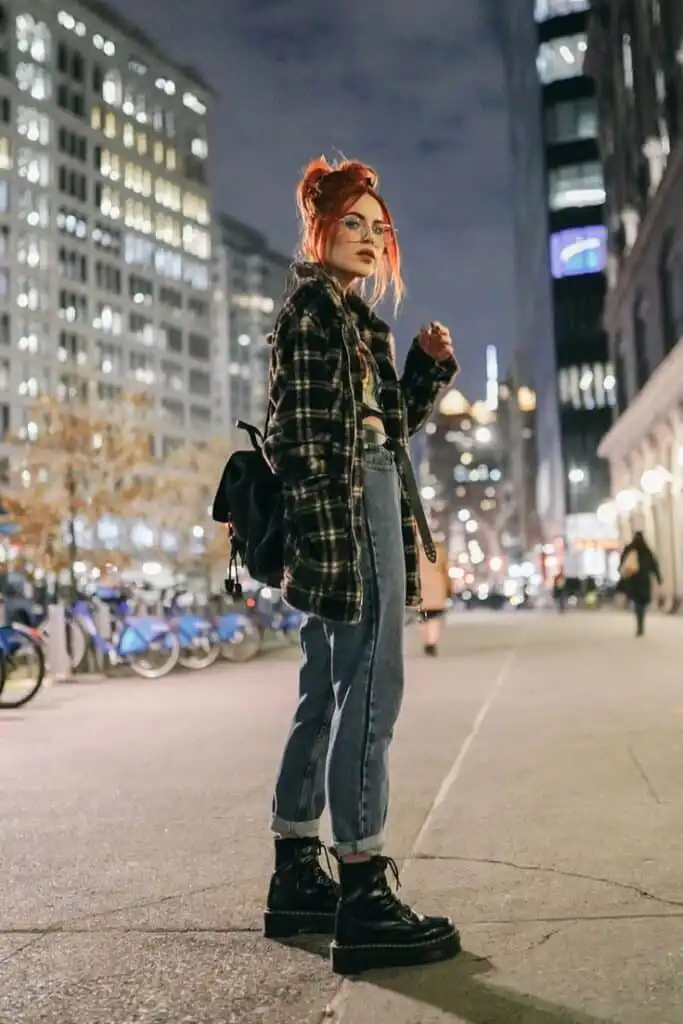 Stylish woman with red hair in a plaid coat and jeans walking through a vibrant city street at night.