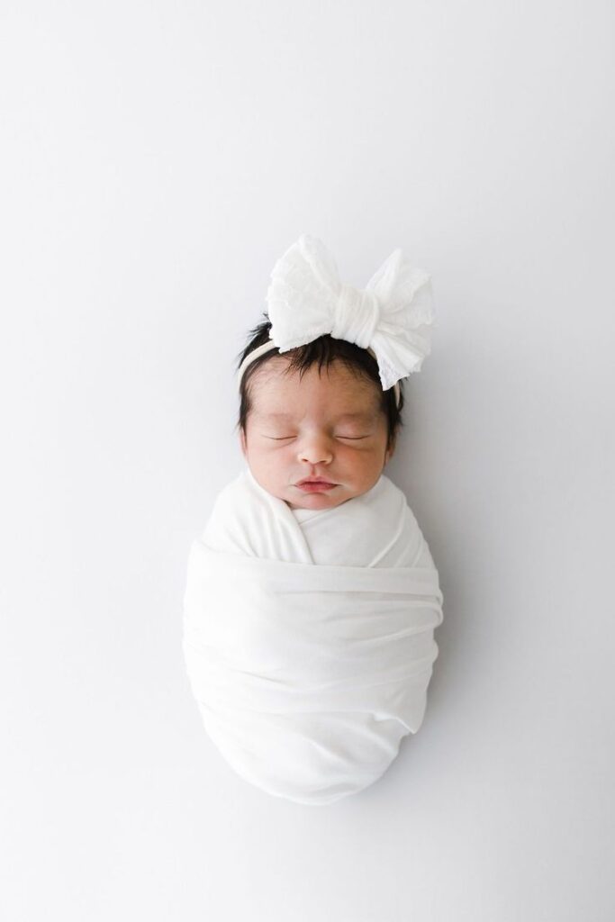How to Use Different Wrapping Techniques for Newborn Photography _ Acapello Photography Newborn baby swaddled in white with a large bow headband, peacefully sleeping on a soft, white background. | Sky Rye Design Newborn baby swaddled in white with a large bow headband, peacefully sleeping on a soft, white background.