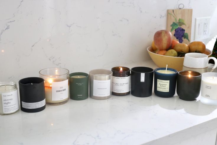 A variety of scented candles in glass jars lined up on a white marble countertop, with a bowl of fruits in the background.