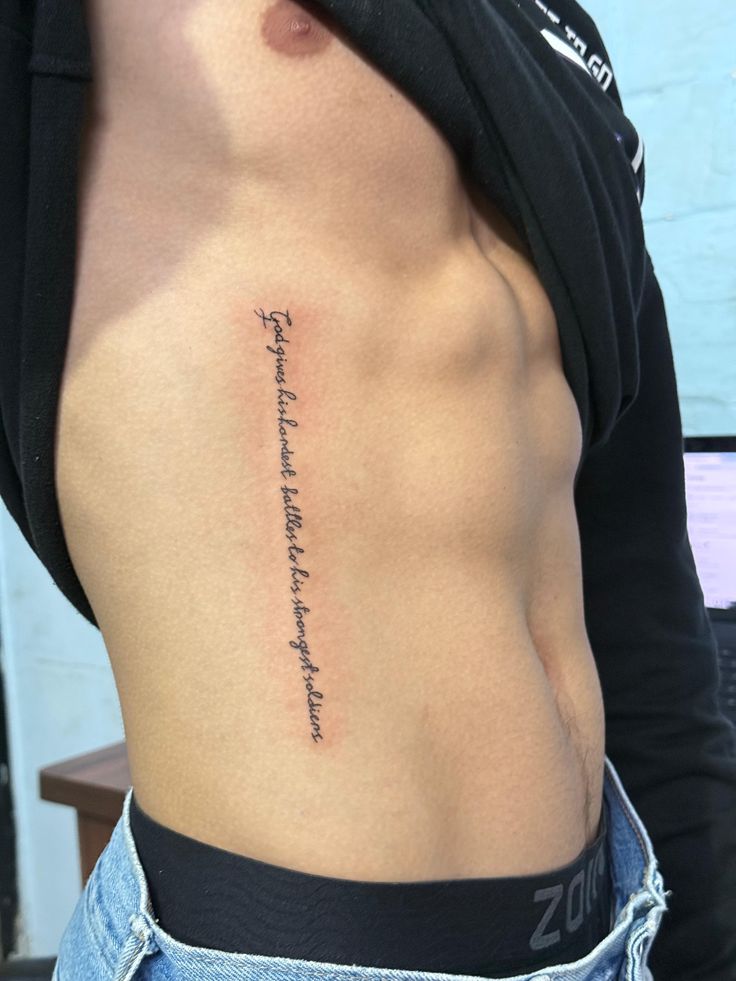 Side view of a person with a script tattoo on their ribcage, wearing jeans and lifting their shirt to reveal the inked message.