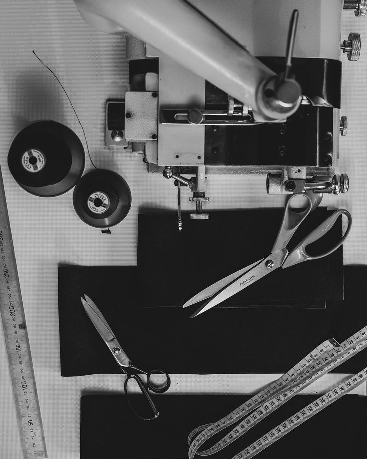 In the quiet of the workshop ready for action___W ciszy pracowni narzędzia gotowe do działania Black and white sewing tools: scissors, thread spools, sewing machine, and measuring tape on a fabric surface, ready for tailoring. | Sky Rye Design Black and white sewing tools: scissors, thread spools, sewing machine, and measuring tape on a fabric surface, ready for tailoring.