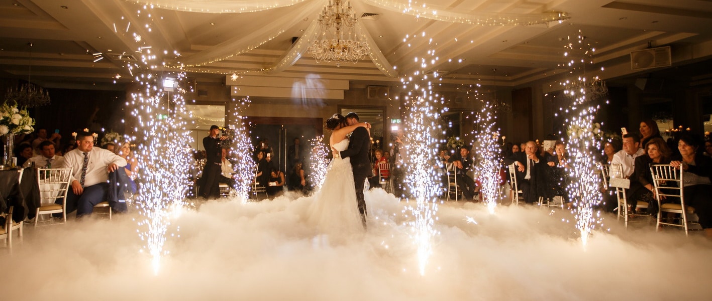 Indoor-Fireworks Bride and groom share first dance with sparklers and fog effects at a wedding reception, surrounded by guests in a grand ballroom. | Sky Rye Design Bride and groom share first dance with sparklers and fog effects at a wedding reception, surrounded by guests in a grand ballroom.