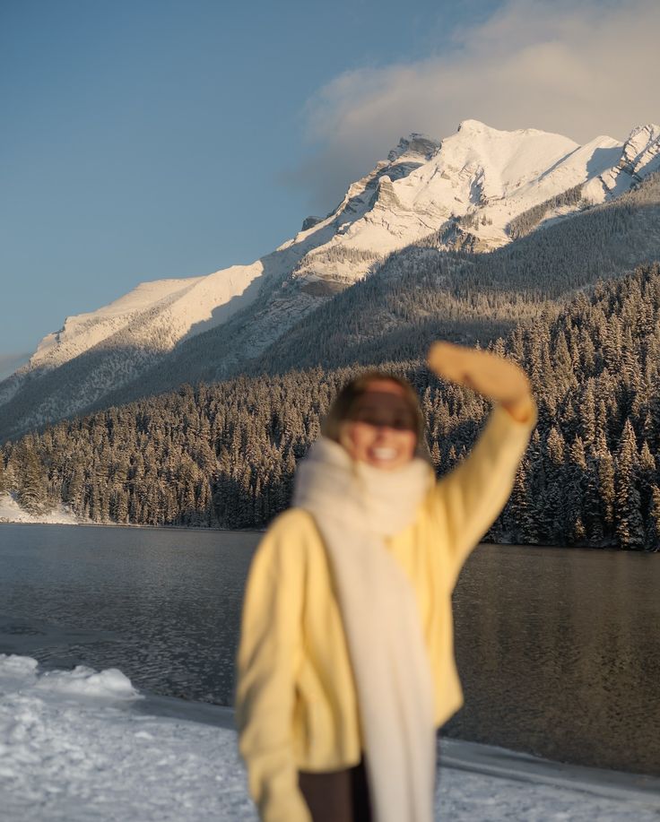 Instagram 1 Person in winter attire smiles and waves by a serene snowy mountain lake under clear skies. | Sky Rye Design Person in winter attire smiles and waves by a serene snowy mountain lake under clear skies.