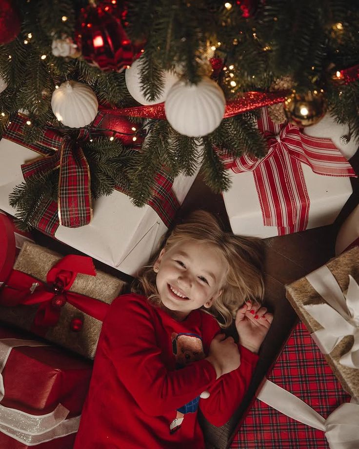 Instagram Smiling child in a red shirt under a decorated Christmas tree surrounded by wrapped gifts with bows and festive ornaments. | Sky Rye Design Smiling child in a red shirt under a decorated Christmas tree surrounded by wrapped gifts with bows and festive ornaments.
